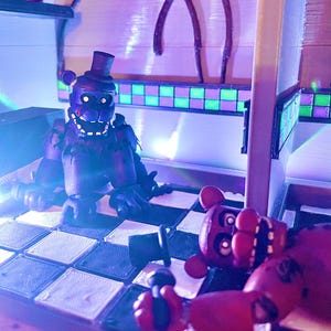 Op de afbeelding: Twee beerachtige figuren met hoge hoeden en microfoons op een zwart-wit geblokte vloer. E&eacute;n figuur is donkerpaars en zit, terwijl de andere roodbruin is en ligt. De sc&egrave;ne wordt verlicht door blauw en paars licht.