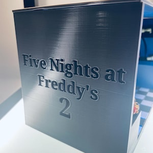 Op de afbeelding: Een donkergrijze, rechthoekige, 3D-geprinte displaybox met de in reli&euml;f aangebrachte tekst "Five Nights at Freddy's 2" op het voorpaneel. De binnenkant van de box toont een blauwachtig geblokte vloer en een klein, kleurrijk figuurtje.