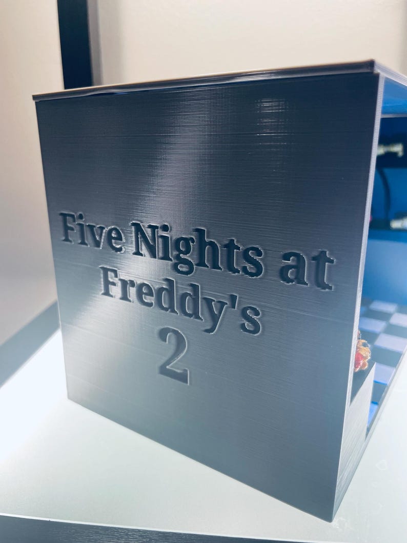 Op de afbeelding: Een donkergrijze, rechthoekige, 3D-geprinte displaybox met de in reli&euml;f aangebrachte tekst "Five Nights at Freddy's 2" op het voorpaneel. De binnenkant van de box toont een blauwachtig geblokte vloer en een klein, kleurrijk figuurtje.