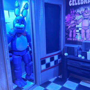 K&ouml;nnte beinhalten: Ein 3D-gedrucktes Diorama mit einer blauen Hasen-Animatronic-Figur, die in einem T&uuml;rrahmen steht. Die Szene umfasst einen schwarz-wei&szlig; karierten Boden, ein Wandplakat mit dem Text CELEBRATE und einen Schreibtisch mit kleinen Ger&auml;ten.