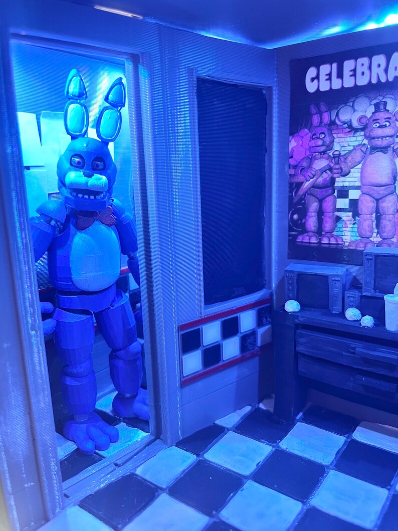 K&ouml;nnte beinhalten: Ein 3D-gedrucktes Diorama mit einer blauen Hasen-Animatronic-Figur, die in einem T&uuml;rrahmen steht. Die Szene umfasst einen schwarz-wei&szlig; karierten Boden, ein Wandplakat mit dem Text CELEBRATE und einen Schreibtisch mit kleinen Ger&auml;ten.
