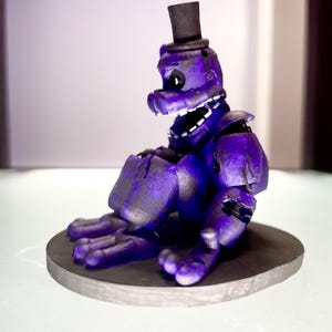 Puede incluir: Figura morada de un personaje con forma de oso con sombrero de copa negro, sentado sobre una base circular gris. La figura tiene una amplia sonrisa con dientes y es una representaci&oacute;n estilizada.