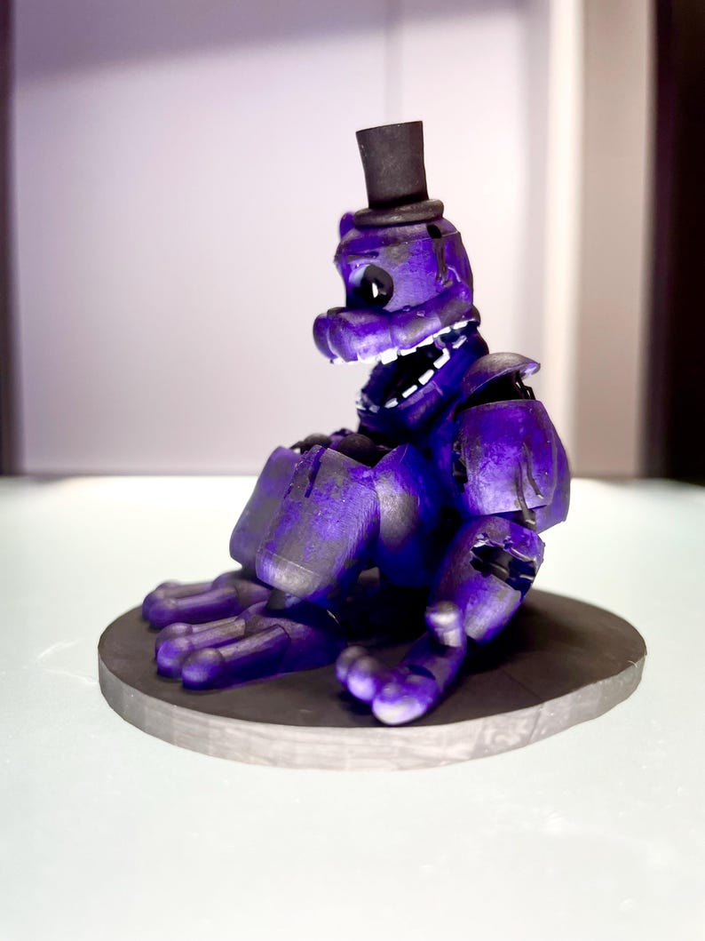 Puede incluir: Figura morada de un personaje con forma de oso con sombrero de copa negro, sentado sobre una base circular gris. La figura tiene una amplia sonrisa con dientes y es una representaci&oacute;n estilizada.