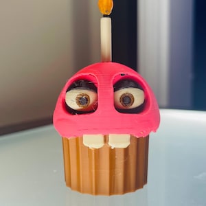 Op de afbeelding: Een 3D-geprinte cupcake-figuur met een felroze bovenkant, een bruine geribbelde basis en twee grote witte ogen met bruine pupillen. Het figuur heeft twee witte tanden die uit het roze glazuur steken en een hoge kaars aan de bovenkant.