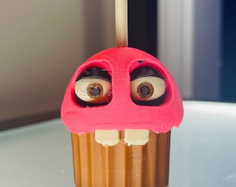 Op de afbeelding: Een 3D-geprinte cupcake-figuur met een felroze bovenkant, een bruine geribbelde basis en twee grote witte ogen met bruine pupillen. Het figuur heeft twee witte tanden die uit het roze glazuur steken en een hoge kaars aan de bovenkant.