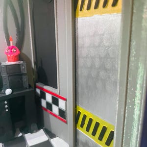 K&ouml;nnte beinhalten: Ein 3D-gedrucktes Diorama mit einer grauen Wand und einer silbernen T&uuml;r, die mit gelben und schwarzen Warnstreifen akzentuiert ist. Eine schwarz-wei&szlig;e Schachbrettbord&uuml;re verl&auml;uft entlang der Wand. Ein kleiner rosa Cupcake steht auf einer schwarzen Konsole.