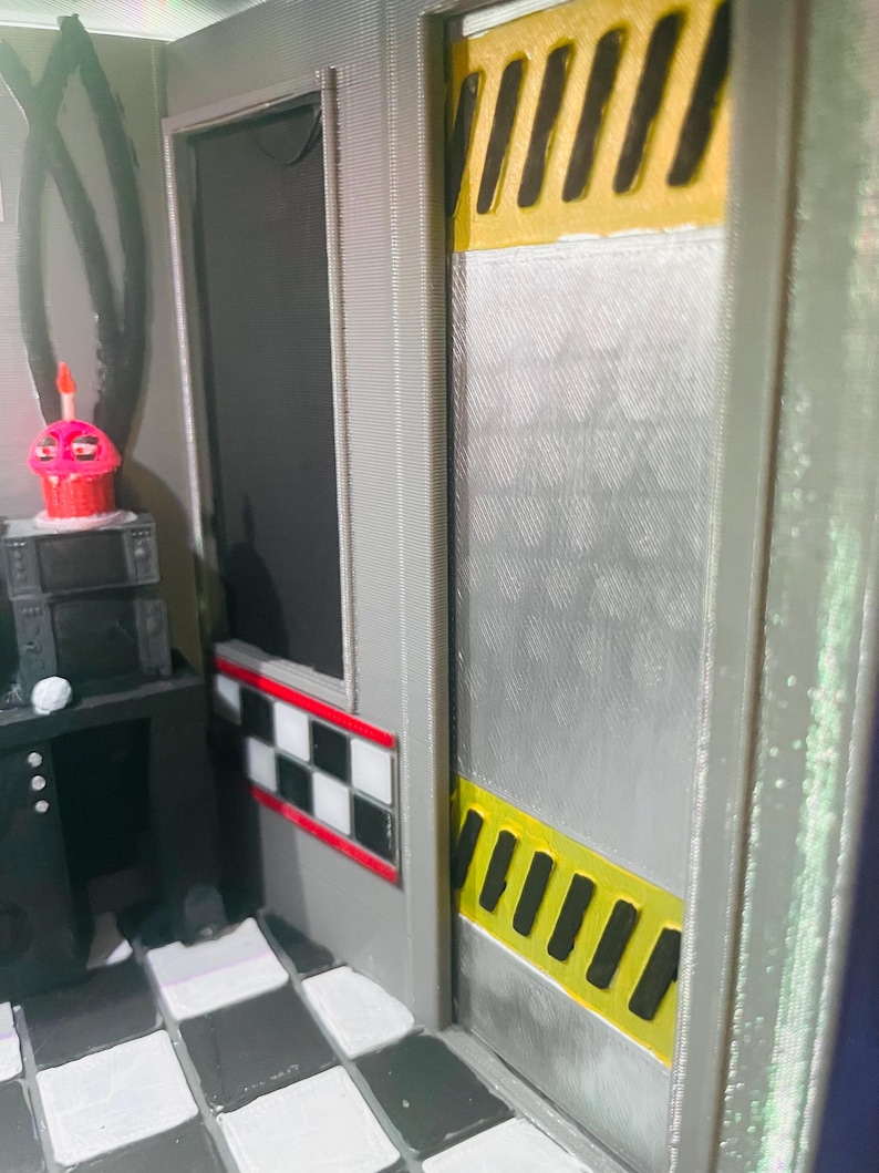 K&ouml;nnte beinhalten: Ein 3D-gedrucktes Diorama mit einer grauen Wand und einer silbernen T&uuml;r, die mit gelben und schwarzen Warnstreifen akzentuiert ist. Eine schwarz-wei&szlig;e Schachbrettbord&uuml;re verl&auml;uft entlang der Wand. Ein kleiner rosa Cupcake steht auf einer schwarzen Konsole.