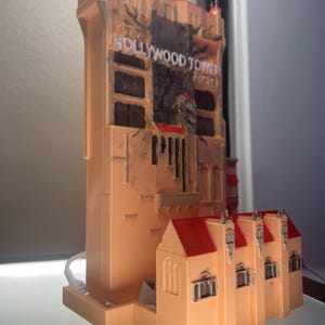 Op de afbeelding: Een gedetailleerd model van het Hollywood Tower Hotel, met een perzikroze buitenkant en rode dakaccenten. Het model heeft de woorden "Hollywood Tower" in witte letters. Het is een replica van de iconische Disney-attractie.