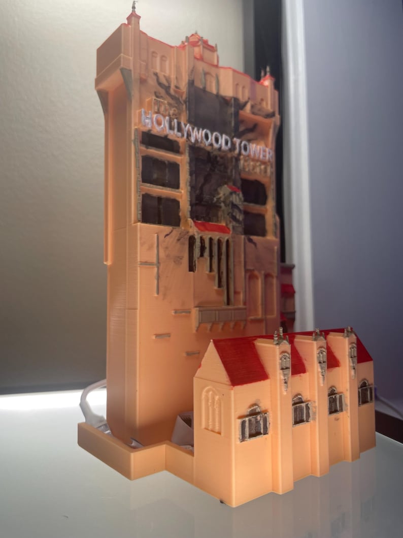 Op de afbeelding: Een gedetailleerd model van het Hollywood Tower Hotel, met een perzikroze buitenkant en rode dakaccenten. Het model heeft de woorden "Hollywood Tower" in witte letters. Het is een replica van de iconische Disney-attractie.