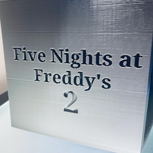 May include: A silver-coloured cube with a smooth, layered texture featuring the black embossed text Five Nights at Freddy's 2 on the front face. The object has a clean, geometric shape with sharp edges and a consistent metallic finish throughout.