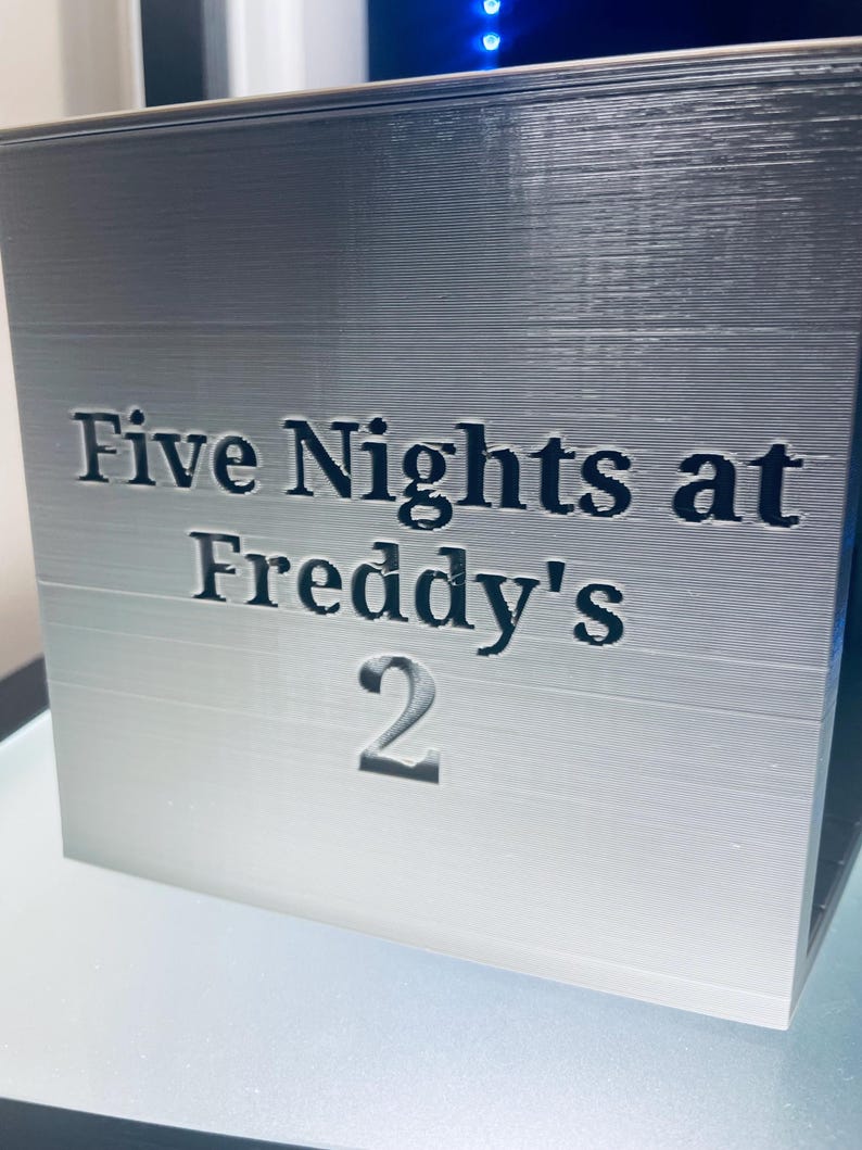 May include: A silver-coloured cube with a smooth, layered texture featuring the black embossed text Five Nights at Freddy's 2 on the front face. The object has a clean, geometric shape with sharp edges and a consistent metallic finish throughout.