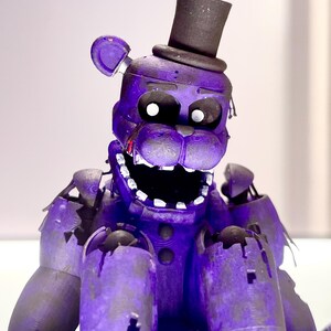 Puede incluir: Una figura morada con forma de oso con un sombrero de copa negro. La figura tiene la boca muy abierta, dientes visibles y una apariencia angustiada. El oso tiene da&ntilde;os y desgaste visibles.