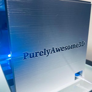 Op de afbeelding: Een metallic zilverkleurige 3D-geprinte kubus met de tekst PurelyAwesome3D in reli&euml;f op het voorpaneel. De kubus heeft een holle binnenkant die wordt verlicht door een felblauw licht en een kleine witte kabel die uit een zijpoort komt.