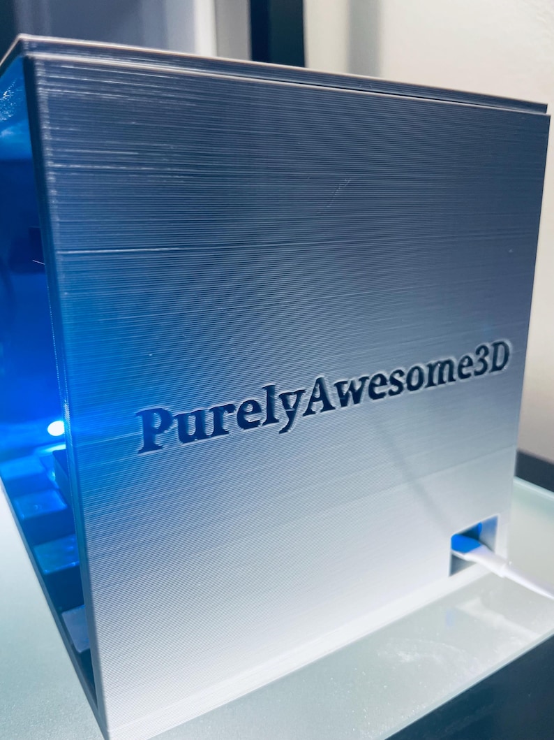 Op de afbeelding: Een metallic zilverkleurige 3D-geprinte kubus met de tekst PurelyAwesome3D in reli&euml;f op het voorpaneel. De kubus heeft een holle binnenkant die wordt verlicht door een felblauw licht en een kleine witte kabel die uit een zijpoort komt.