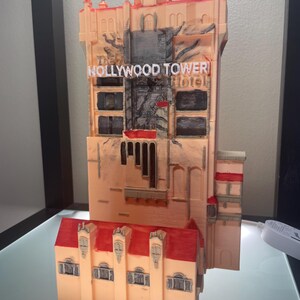 Op de afbeelding: Een 3D-geprint model van het Hollywood Tower Hotel, met een perzikkleurige buitenkant, rode accenten en een rood dak. Het model bevat de naam van het hotel in witte letters en gedetailleerde architectonische elementen.