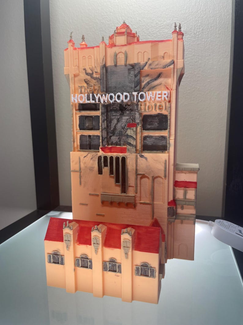 Op de afbeelding: Een 3D-geprint model van het Hollywood Tower Hotel, met een perzikkleurige buitenkant, rode accenten en een rood dak. Het model bevat de naam van het hotel in witte letters en gedetailleerde architectonische elementen.