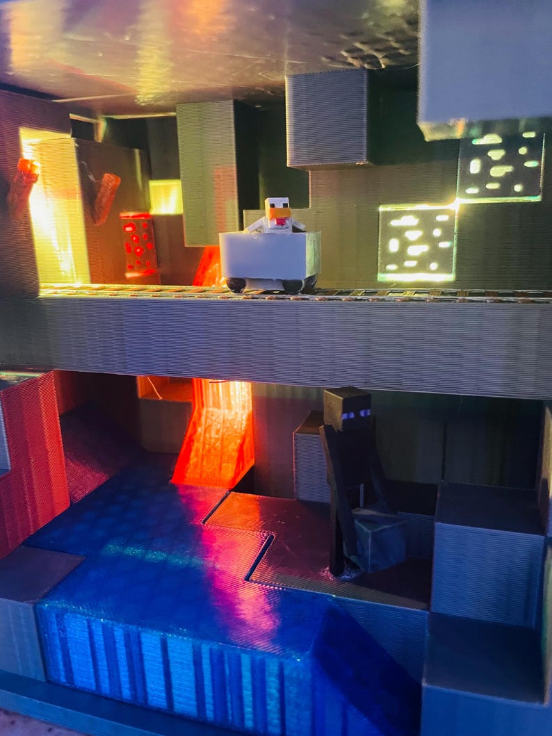 Puede incluir: Un diorama impreso en 3D con tem&aacute;tica de Minecraft con un pollo montando en un carro de mina, una cascada de lava y una figura negra de pie en una plataforma.