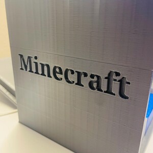 Puede incluir: Una caja gris impresa en 3D con la palabra "Minecraft" impresa en negro en la parte delantera.