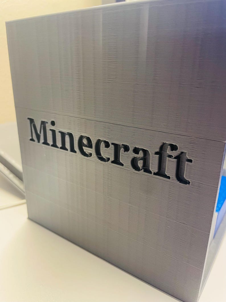 Puede incluir: Una caja gris impresa en 3D con la palabra "Minecraft" impresa en negro en la parte delantera.