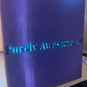 K&ouml;nnte beinhalten: Ein rechteckiges, violettes 3D-gedrucktes Objekt mit glatter Oberfl&auml;che und horizontalen Schichtlinien. Der Text "Purely Awesome 3D" ist auf der Vorderseite eingepr&auml;gt. Ein helles blaues Licht strahlt von der Oberkante und beleuchtet die Schrift.