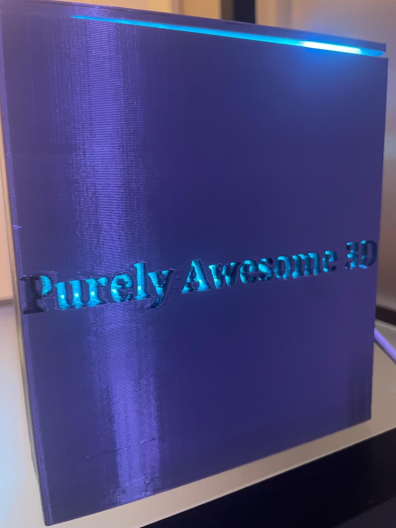 K&ouml;nnte beinhalten: Ein rechteckiges, violettes 3D-gedrucktes Objekt mit glatter Oberfl&auml;che und horizontalen Schichtlinien. Der Text "Purely Awesome 3D" ist auf der Vorderseite eingepr&auml;gt. Ein helles blaues Licht strahlt von der Oberkante und beleuchtet die Schrift.