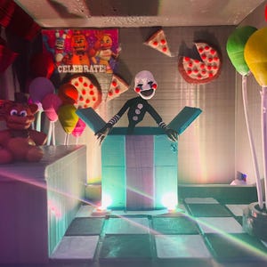 May include: A diorama featuring a puppet character emerging from a teal box, surrounded by balloons, pizza slices, and a brown teddy bear. A banner that says "CELEBRATE!" is visible in the background. The scene is lit with colorful lights.