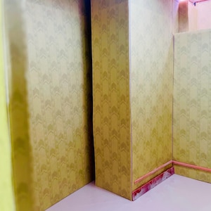 May include: A miniature room interior featuring walls covered in a light yellow wallpaper with a repeating vertical chevron pattern. The structure includes a central pillar, a light pink floor, and a thin pink trim along the base of the walls.