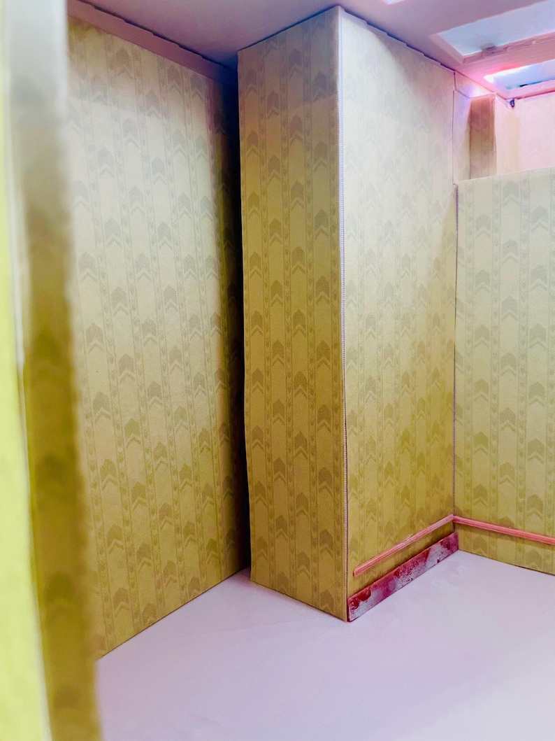 May include: A miniature room interior featuring walls covered in a light yellow wallpaper with a repeating vertical chevron pattern. The structure includes a central pillar, a light pink floor, and a thin pink trim along the base of the walls.
