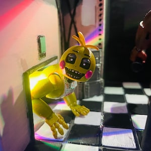 May include: A yellow animatronic chicken figure with pink cheeks and a white bib featuring colourful confetti dots. The figure is emerging from a doorway onto a black and white checkered floor, illuminated by a bright yellow light from behind.