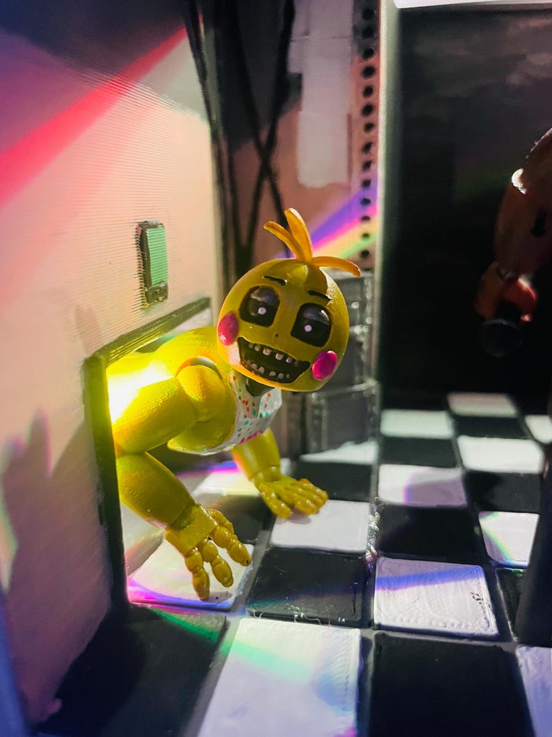 May include: A yellow animatronic chicken figure with pink cheeks and a white bib featuring colourful confetti dots. The figure is emerging from a doorway onto a black and white checkered floor, illuminated by a bright yellow light from behind.