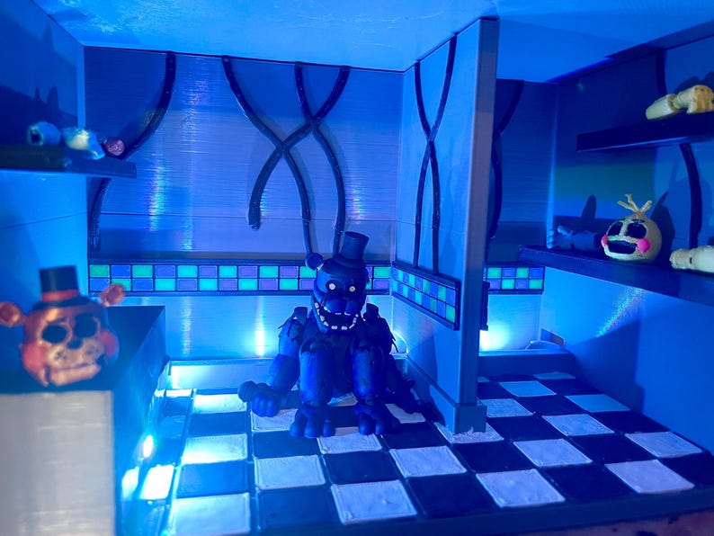 Op de afbeelding: Een miniatuur diorama met een donkerblauwe beer animatronic figuur zittend op een zwart-wit geblokte vloer. De sc&egrave;ne bevat een berenkop op een toonbank, een gele vogelkop op een plank en blauwe verlichting door de hele kamer.