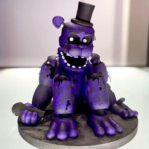 Op de afbeelding: Een paars berenfiguur met een zwarte hoge hoed en microfoon, met tekenen van slijtage. Het figuur staat op een donkergrijze, cirkelvormige basis. De ogen zijn wit.