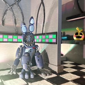 Op de afbeelding: Een blauwe animatronic konijnfiguur met een beschadigd gezicht en blootliggende mechanische onderdelen, zittend op een zwart-wit geblokte vloer. Het figuur heeft een rode vlinderdas en staat in een kamer met grijze muren en groene tegels.