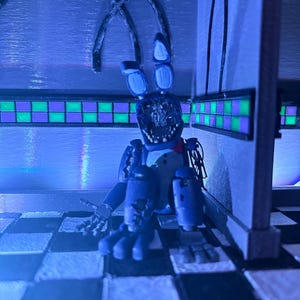 Op de afbeelding: Een blauwe animatronic konijnfiguur met een beschadigd gezicht, blootliggende mechanische onderdelen en lange oren, zittend op een zwart-wit geblokte vloer. Het figuur heeft een rode vlinderdas en staat in een hoek met groen-paarse tegels.