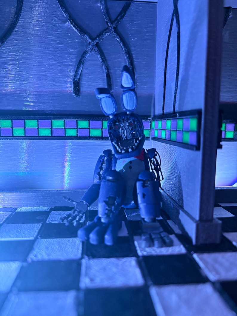 Op de afbeelding: Een blauwe animatronic konijnfiguur met een beschadigd gezicht, blootliggende mechanische onderdelen en lange oren, zittend op een zwart-wit geblokte vloer. Het figuur heeft een rode vlinderdas en staat in een hoek met groen-paarse tegels.