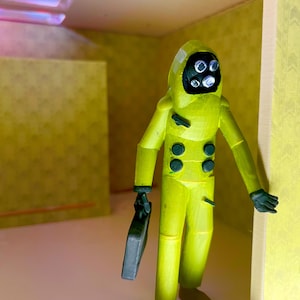May include: A yellow hazmat suit figurine with a black face mask featuring four circular eyes. The figure has dark green buttons on the chest and holds a small black rectangular briefcase in one hand. It stands within a yellow-walled room.