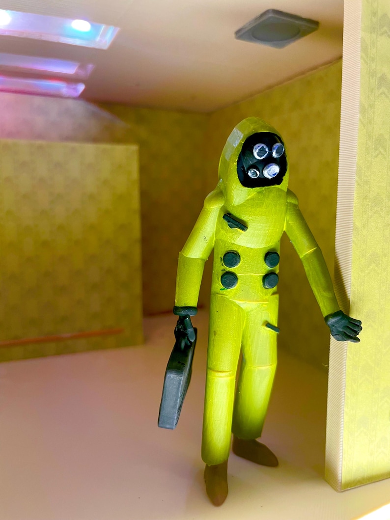 May include: A yellow hazmat suit figurine with a black face mask featuring four circular eyes. The figure has dark green buttons on the chest and holds a small black rectangular briefcase in one hand. It stands within a yellow-walled room.