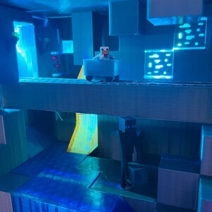 Puede incluir: Un diorama de Minecraft de color azul y gris con una peque&ntilde;a figura de una oveja conduciendo un carro azul. El diorama presenta una cascada y una figura de un personaje vestido de negro.