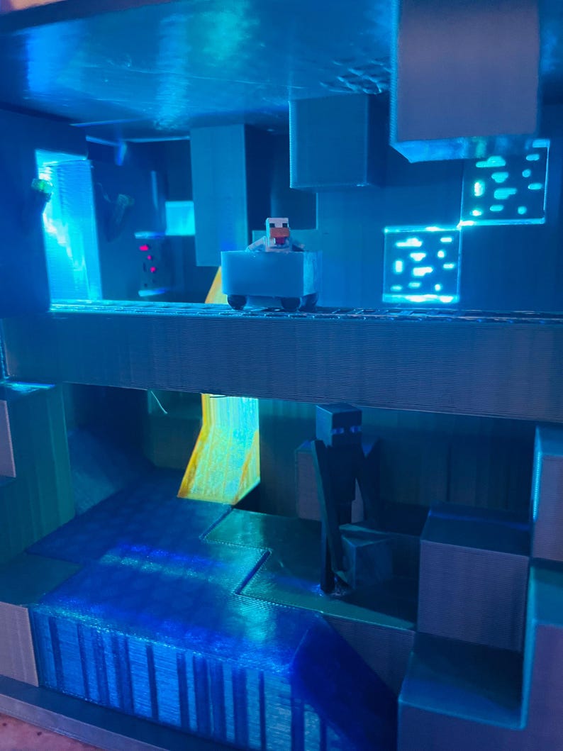 Puede incluir: Un diorama de Minecraft de color azul y gris con una peque&ntilde;a figura de una oveja conduciendo un carro azul. El diorama presenta una cascada y una figura de un personaje vestido de negro.