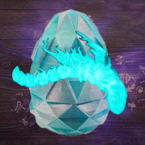 May include: A light blue, geometric, faceted egg-shaped object featuring a bright, glowing cyan silhouette of a creature with multiple legs and a long, curved tail positioned horizontally across the centre of the faceted surface.