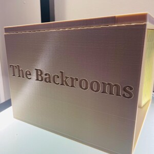 May include: A beige, rectangular, 3D-printed box with the text "The Backrooms" embossed on the front side. The object features a smooth, layered surface texture characteristic of additive manufacturing, with a flat top and a solid, blocky construction.