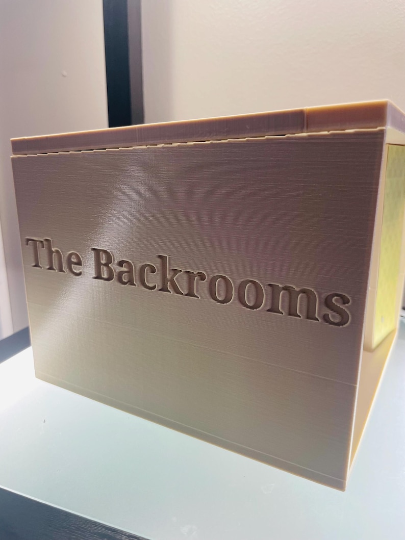 May include: A beige, rectangular, 3D-printed box with the text "The Backrooms" embossed on the front side. The object features a smooth, layered surface texture characteristic of additive manufacturing, with a flat top and a solid, blocky construction.