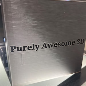 May include: A metallic silver, rectangular 3D printed object with a smooth, layered finish. The front face features the text "Purely Awesome 3D" embossed in a dark, bold font. The surface displays horizontal lines characteristic of the 3D printing process.