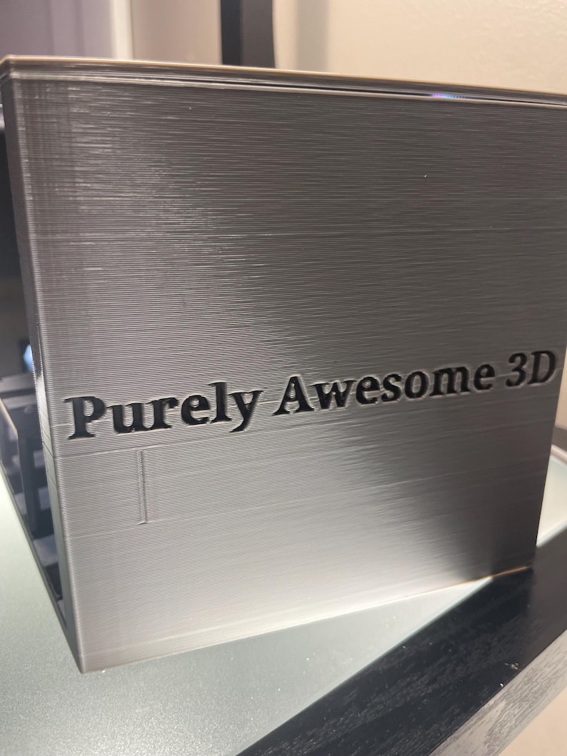 May include: A metallic silver, rectangular 3D printed object with a smooth, layered finish. The front face features the text "Purely Awesome 3D" embossed in a dark, bold font. The surface displays horizontal lines characteristic of the 3D printing process.