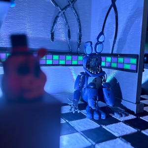 Op de afbeelding: Een diorama met een blauwe animatronic konijnenfiguur met een beschadigd gezicht, zittend op een zwart-wit geblokte vloer. De sc&egrave;ne bevat een muur met een groen en paars betegelde rand en een wazig figuur op de voorgrond onder blauw licht.