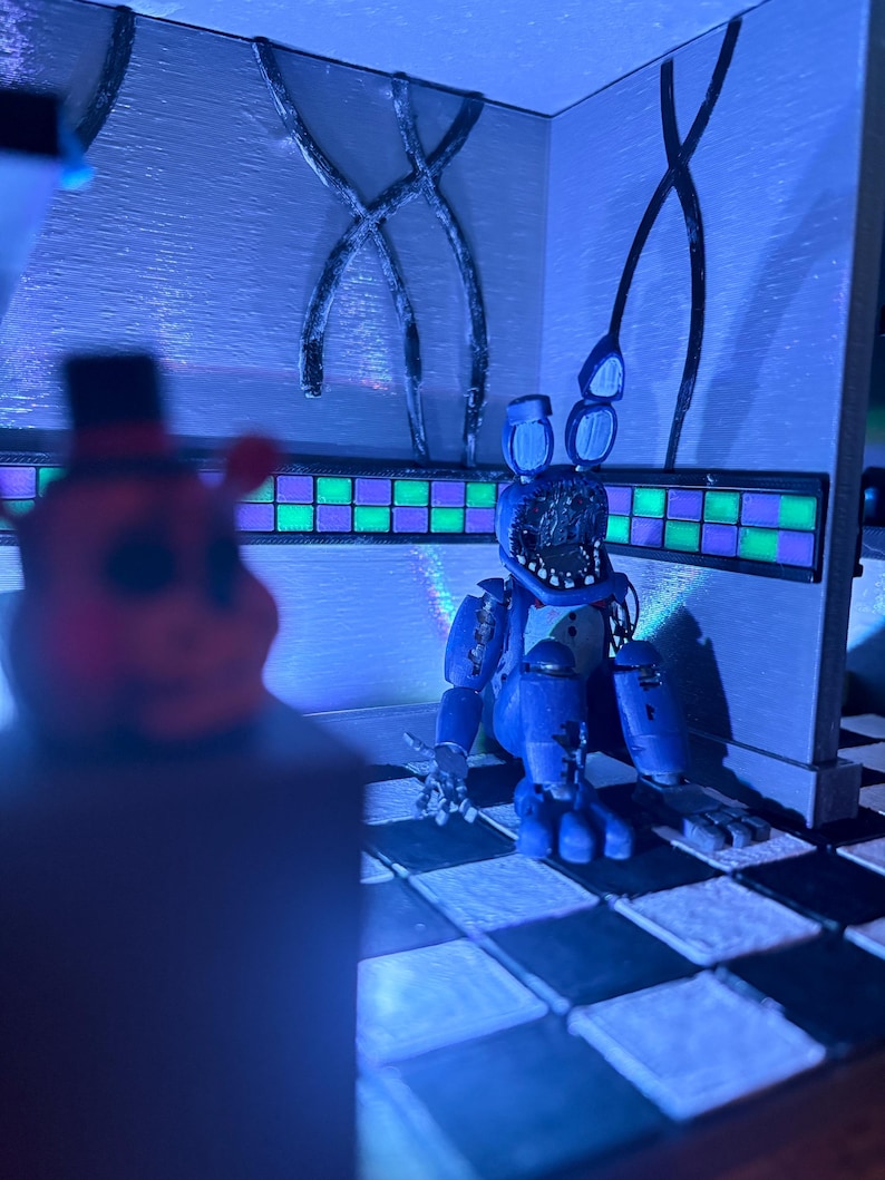 Op de afbeelding: Een diorama met een blauwe animatronic konijnenfiguur met een beschadigd gezicht, zittend op een zwart-wit geblokte vloer. De sc&egrave;ne bevat een muur met een groen en paars betegelde rand en een wazig figuur op de voorgrond onder blauw licht.
