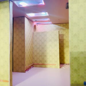 May include: A miniature room interior featuring light yellow walls with a repeating geometric arrow pattern. The space includes a smooth, light-colored floor and a ceiling equipped with three recessed rectangular lights emitting a soft glow.