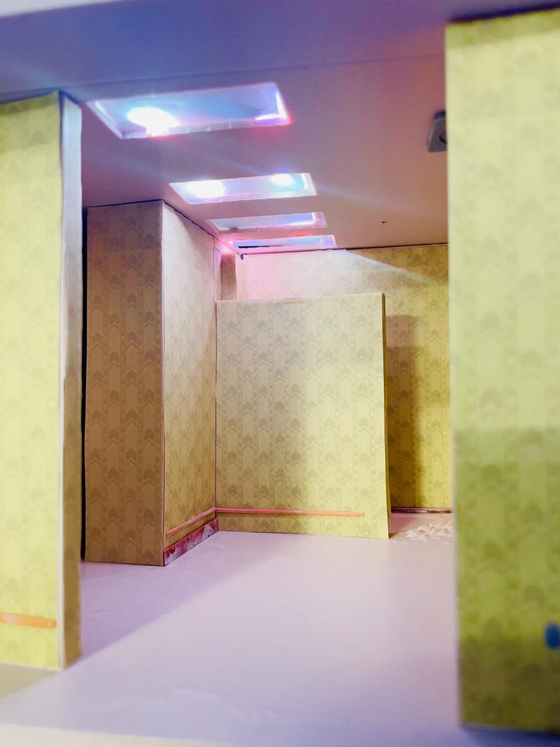 May include: A miniature room interior featuring light yellow walls with a repeating geometric arrow pattern. The space includes a smooth, light-colored floor and a ceiling equipped with three recessed rectangular lights emitting a soft glow.