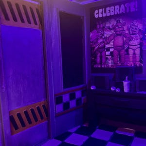 K&ouml;nnte beinhalten: Ein Miniatur-Diorama eines Sicherheitsb&uuml;ros mit schwarz-wei&szlig; kariertem Boden, einer Wand mit einem Poster von drei Animatronic-Figuren und dem Text CELEBRATE!, einer dunklen T&uuml;r mit orangefarbenen L&uuml;ftungsschlitzen und violetter Beleuchtung.
