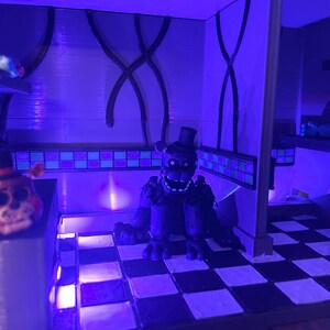 Op de afbeelding: Een diorama met een donker, verweerd berenfiguur met een hoge hoed op een zwart-wit geblokte vloer. Een kleinere berenkop met een hoge hoed staat op een richel aan de linkerkant en een gele vogelachtige kop staat op een plank aan de rechterkant.